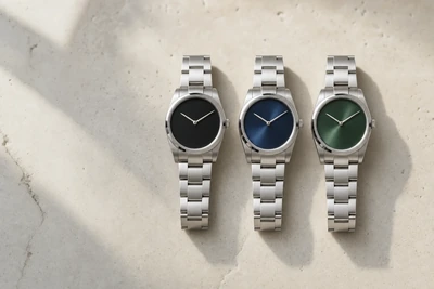 Três relógios de mostrador preto, azul e verde lado a lado sobre pedra clara