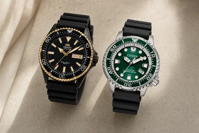 Orient Kamasu preto e dourado ao lado de um Citizen Promaster verde sobre pedra clara