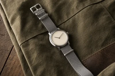 Relógio de aço com pulseira de tecido cinza passando por baixo da caixa, sobre lona verde